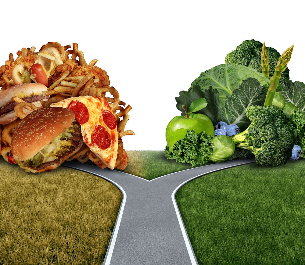 A road, with one branch leading to a mountain of pizza, fries, and hamburgers and the other leading to a pile of broccoli, apples, and asparagus, representing nutrition counseling choices